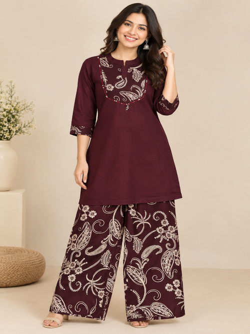 A woman modeling a three-piece maroon kurta set with white floral embroidery, featuring a straight kurta, embroidered yoke, and wide-leg pants, set against a neutral beige background.