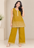 A woman models a mustard yellow two-piece kurta set with white embroidery, featuring a sleeveless tunic top and matching wide-leg pants, set against a neutral beige background.